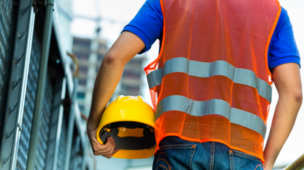 Trabalhador com colete refletivo segura capacete de segurança em área industrial, simbolizando afastamento, desligamento ou mudança de função.