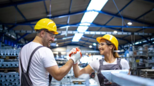 Dois trabalhadores usando capacetes de segurança e EPIs se cumprimentam em ambiente industrial, simbolizando trabalho em equipe e segurança no trabalho.