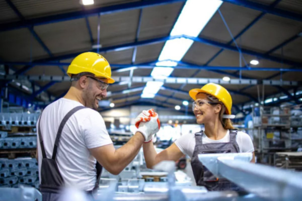 Dois trabalhadores usando capacetes de segurança e EPIs se cumprimentam em ambiente industrial, simbolizando trabalho em equipe e segurança no trabalho.