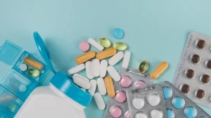 Comprimidos e cápsulas de diferentes cores e formatos espalhados sobre um fundo azul, ao lado de cartelas de medicamentos e um organizador de remédios aberto