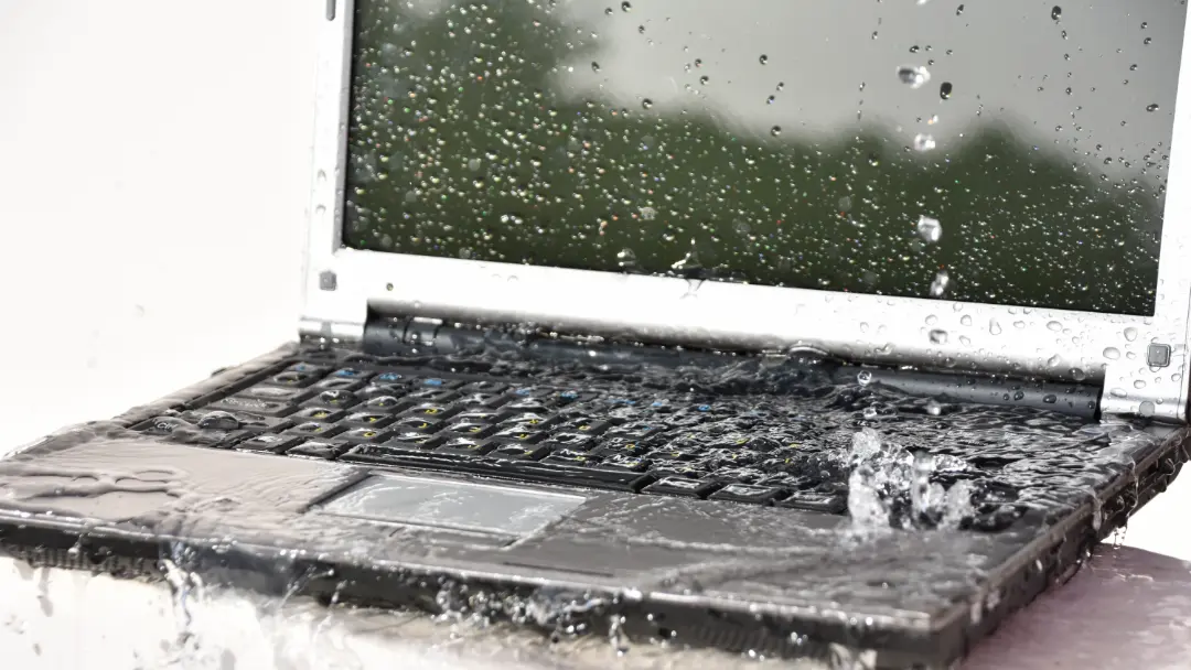 Notebook aberto sendo atingido por respingos de água sobre o teclado e a tela, ilustrando risco de danos por líquidos em equipamentos eletrônicos