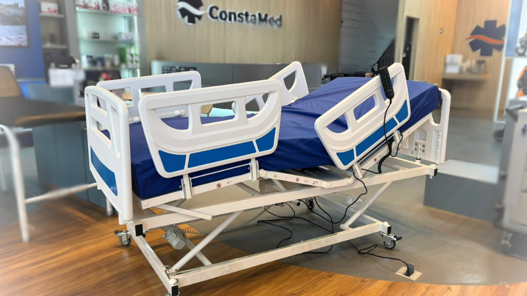 Cama hospitalar elétrica com elevação de encosto e pernas, grades laterais e controle remoto, exposta em showroom de equipamentos médicos.