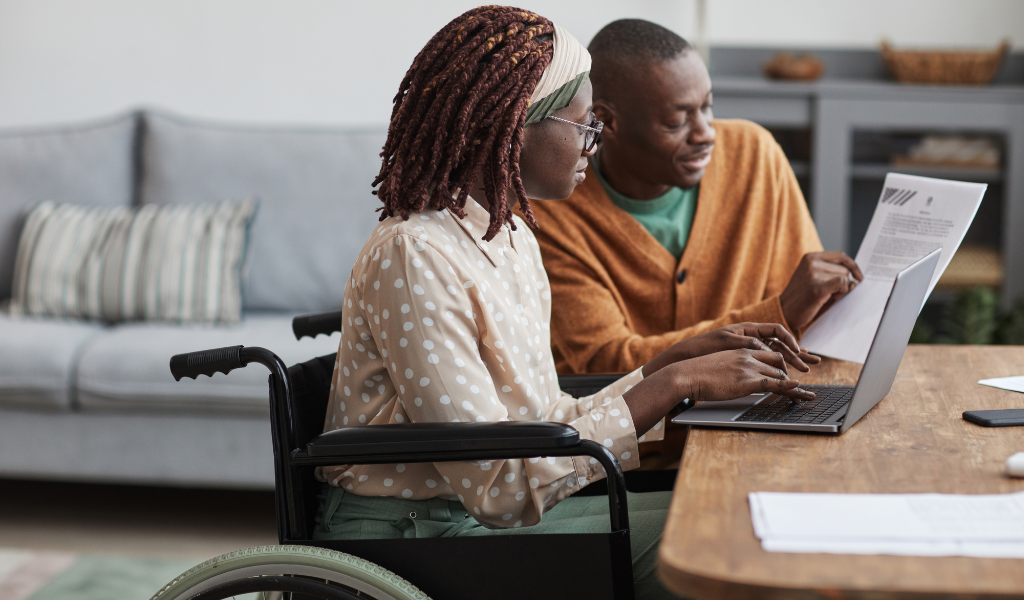 Imagem mostra mulher em cadeira de rodas utilizando notebook com apoio de acompanhante. Representa acessibilidade, inclusão digital e autonomia para pessoa com deficiência no ambiente domiciliar.