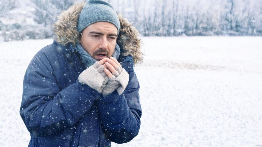 Homem exposto ao frio intenso em ambiente nevado apresenta sinais de hipotermia, como tremores e dificuldade para se aquecer. A imagem ilustra os riscos da baixa temperatura corporal e a importância da prevenção e atendimento rápido.
