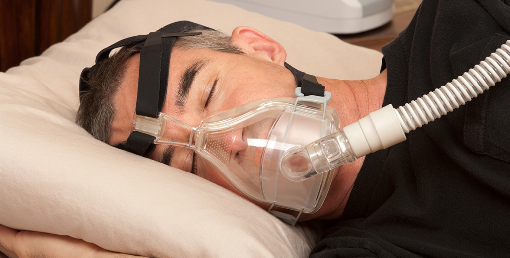 Homem dormindo de lado usando máscara CPAP conectada a equipamento de ventilação. A máscara cobre nariz e boca e está presa por tiras ajustáveis, auxiliando no tratamento da apneia do sono durante a noite.