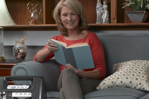 Mulher idosa sentada no sofá lendo um livro enquanto utiliza oxigenoterapia domiciliar por cânula nasal. A imagem representa qualidade de vida, bem-estar e tratamento respiratório em casa.