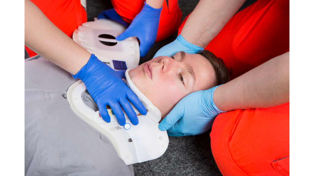 Profissionais de emergência realizam imobilização cervical em paciente deitado no chão, utilizando colar cervical rígido e luvas descartáveis. Procedimento aplicado em casos de trauma para estabilizar a coluna e prevenir lesões.