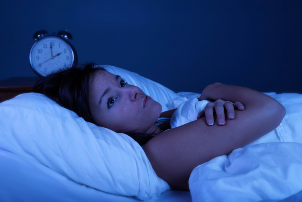 Imagem mostra mulher acordada na cama durante a noite, com expressão de preocupação e relógio ao fundo. Representa insônia, dificuldade para dormir e distúrbios do sono que afetam a qualidade de vida.