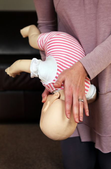 Pessoa demonstra técnica de desengasgamento em bebê utilizando boneco, segurando-o de cabeça para baixo para aplicação de tapotagens nas costas. Imagem ilustrativa de primeiros socorros para obstrução das vias aéreas em lactentes.