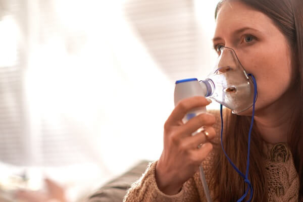 Mulher utilizando máscara de nebulização conectada a aparelho inalador em ambiente interno. A imagem representa tratamento respiratório para asma, bronquite ou outras condições pulmonares.