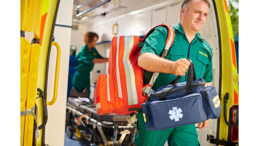 Paramédico uniformizado sai de ambulância carregando mochila e bolsa de atendimento médico de emergência. Ao fundo, maca e equipamentos internos do veículo indicam atendimento pré-hospitalar em ação.