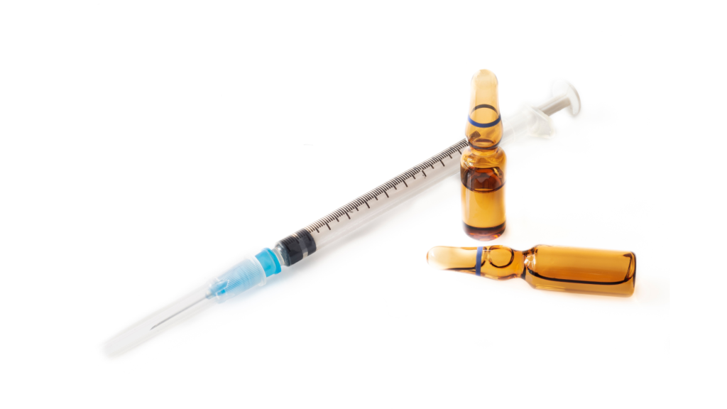 Seringa descartável com agulha ao lado de ampolas de vidro âmbar contendo medicamento injetável. Itens utilizados para aplicação de fármacos por via intravenosa ou intramuscular em ambientes hospitalares e clínicos.