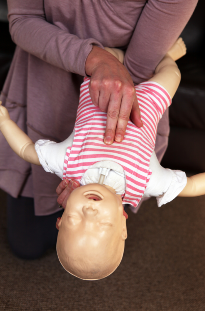 Demonstração de técnica de primeiros socorros para desengasgo em bebê com uso de boneco, segurado de cabeça para baixo enquanto são aplicadas tapotagens nas costas. Procedimento indicado para desobstrução das vias aéreas em lactentes.