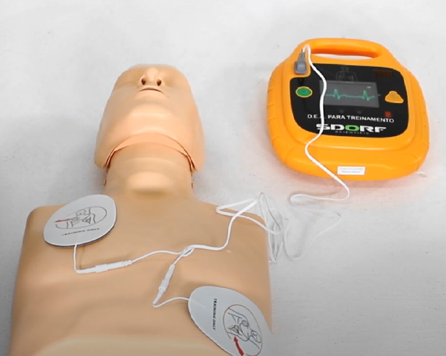 Imagem mostra o uso do DEA (desfibrilador externo automático) na ressuscitação cardiopulmonar (RCP) em adulto, com posicionamento correto dos eletrodos em manequim de treinamento para parada cardiorrespiratória.