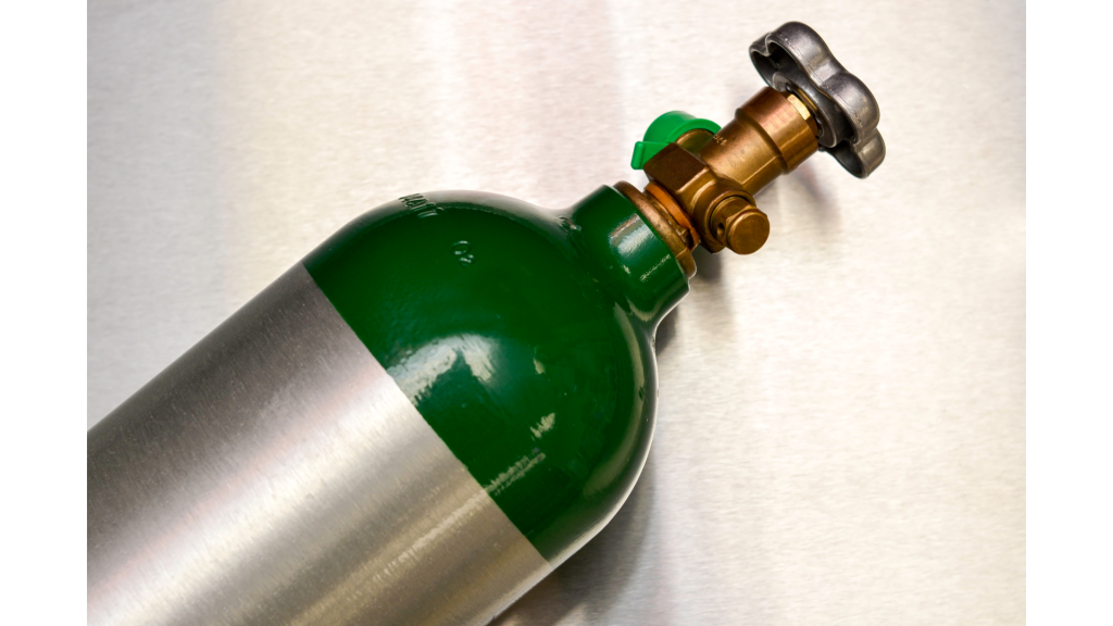 Detalhe da válvula de um cilindro de oxigênio medicinal com parte superior verde e corpo metálico. A imagem mostra o registro e a saída de conexão, componentes responsáveis pelo controle do fluxo de oxigênio.