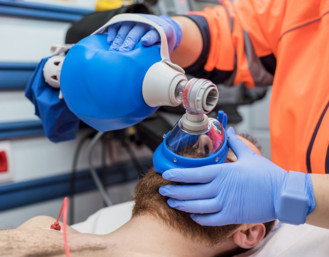 Imagem de profissional de saúde realizando ventilação manual com AMBU (bolsa-válvula-máscara) em paciente durante atendimento de emergência. Procedimento essencial no suporte básico e avançado de vida em casos de parada ou insuficiência respiratória.