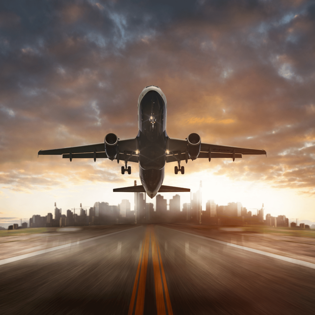 Avião comercial decolando de uma pista de aeroporto com a silhueta de uma cidade ao fundo durante o pôr do sol. A imagem mostra a aeronave vista de frente no momento da subida, com céu dramático e luz dourada iluminando a cena.