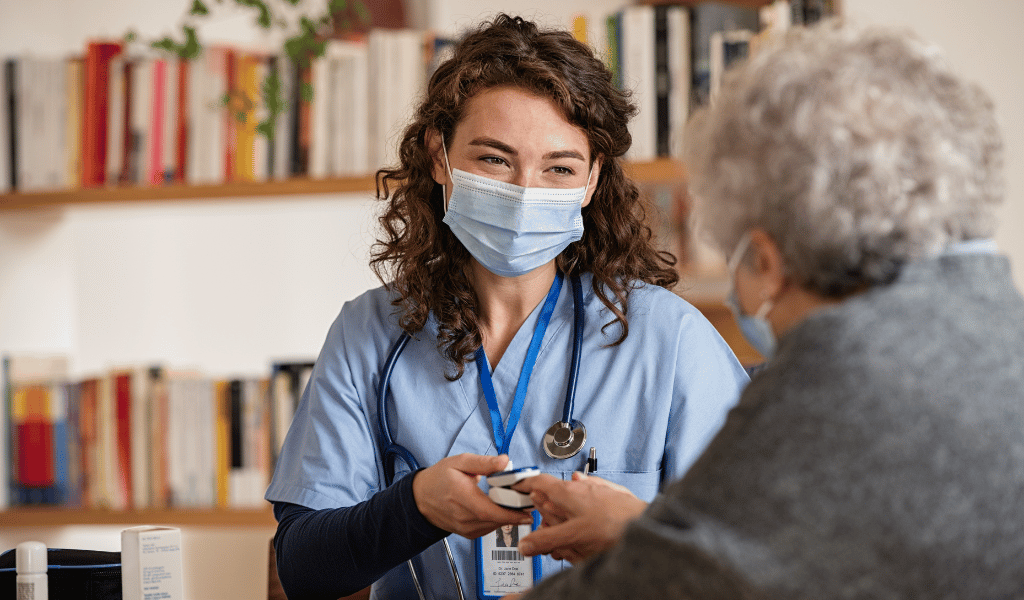 Profissional de saúde usando máscara e estetoscópio atende uma idosa durante consulta. A profissional segura um oxímetro de dedo enquanto conversa com a paciente, em ambiente interno com estante de livros ao fundo.