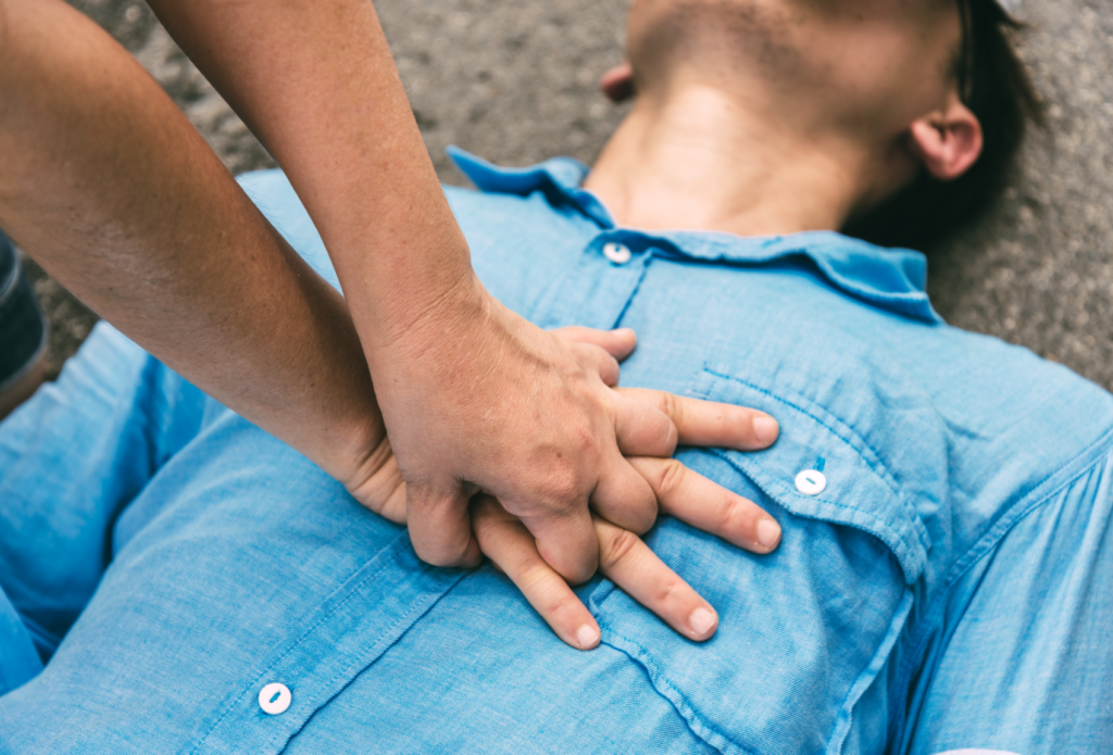 Pessoa realiza compressões torácicas em homem deitado no chão, aplicando técnica de RCP. A imagem representa atendimento de primeiros socorros em caso de parada cardiorrespiratória em ambiente externo.
