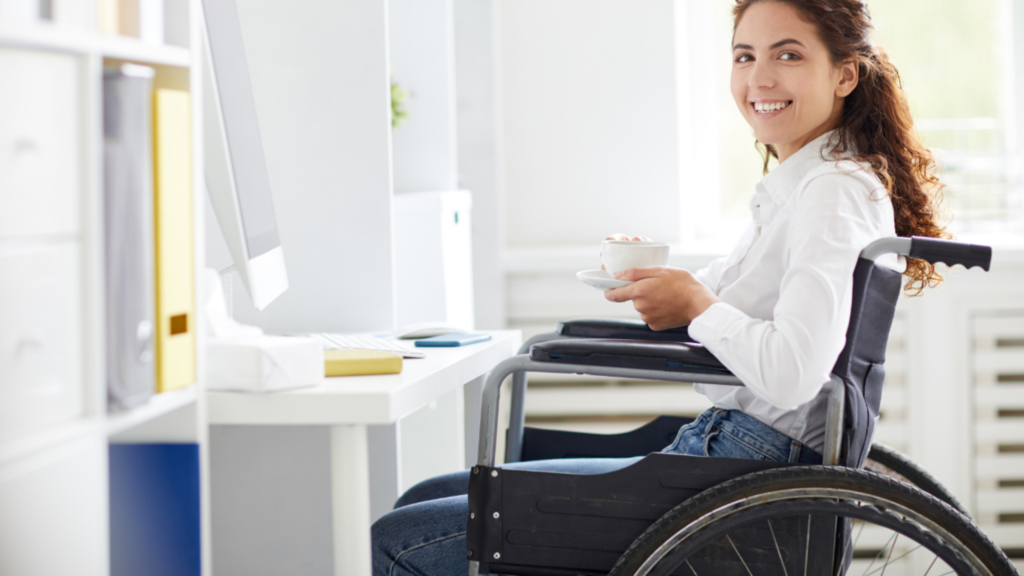 Mulher sorridente em cadeira de rodas trabalhando em escritório moderno. Ela segura uma xícara de café enquanto está sentada diante do computador, em ambiente claro e organizado, representando inclusão e acessibilidade no trabalho.