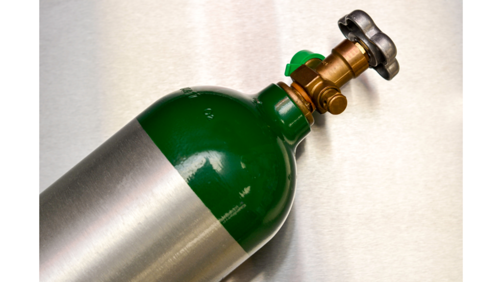 Detalhe da válvula de um cilindro de oxigênio medicinal com parte superior verde e corpo metálico. A imagem mostra o registro e a saída de conexão, componentes responsáveis pelo controle do fluxo de oxigênio.